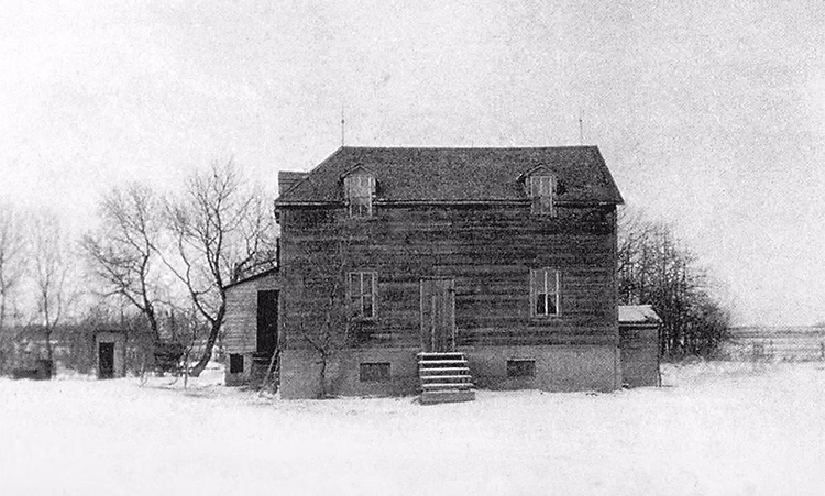 Photo en noir et blanc d’une maison en bois en hiver sur un terrain enneigé.