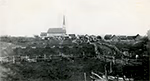 Vue ancienne de la ville de Jonquière en 1876, montrant un paysage rural avec clôtures en bois, maisons dispersées et une grande église à clocher au centre.