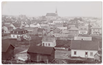 Vue ancienne de la ville de Jonquière, montrant un regroupement de maisons en bois et une grande église à clocher en arrière-plan, dans un paysage urbain de la fin du XIXe ou du début du XXe siècle.