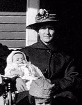 Portrait ancien en noir et blanc d’une personne assise tenant un bébé emmailloté, avec vêtements d’époque et chapeau élaboré, pris en 1919.
