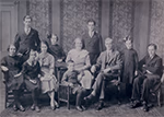 Portrait en noir et blanc d’un groupe de dix personnes posant dans un intérieur ancien avec papier peint à motifs et mobilier en bois, évoquant le début du XXe siècle.