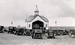 Photo en noir et blanc d’un petit bâtiment religieux, le premier oratoire du Sacré-Cœur de Lebret, avec des voitures garées devant.